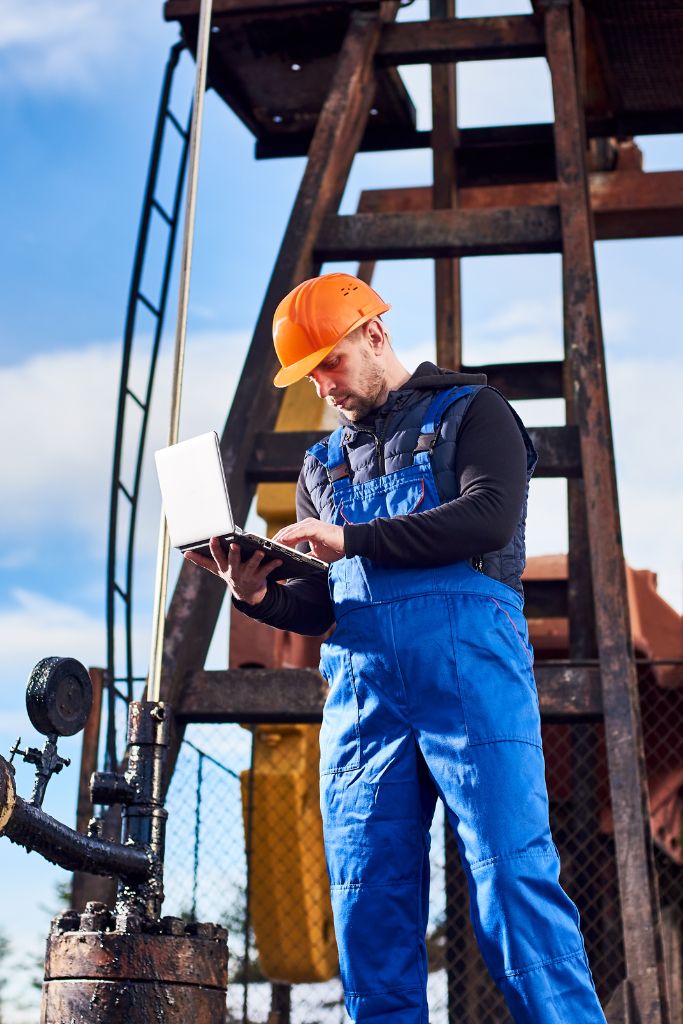 A supervisor in a jumpsuit using a laptop at an oil and gas site, integrating IT solutions into field operations.
