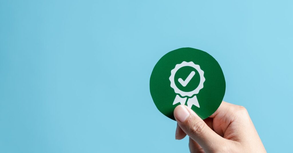 A hand holding a certification badge with a checkmark, symbolizing ISO 27001 compliance and the importance of information security standards.